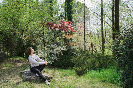 happy caucasian young adult woman sitting on stone in wild wood or national park, sun and forest bathing shinrin yoku concept, horizontal lifestyle outdoors stock photo image
