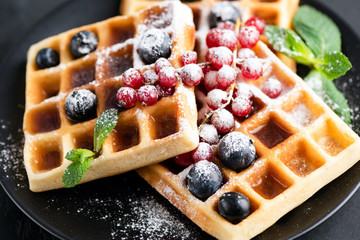 Square belgian waffles with berries and icing sugar on a black plate. Tasty sweet sugary waffles
