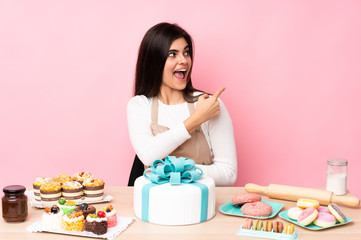 Pastry chef with a big cake in a table over isolated pink background surprised and pointing side