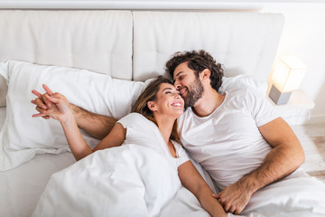 Happy couple is lying in bed together. Enjoying the company of each other.Happy young couple hugging and smiling while lying on the bed in a bedroom at home.