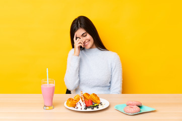 Young woman eating waffles and milkshake in a table over isolated yellow background laughing