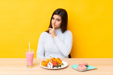 Young woman eating waffles and milkshake in a table over isolated yellow background doing silence gesture