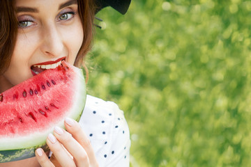 Pretty girl in black hat is eating slice of fresh watermelon in nature. Young woman eats piece of...