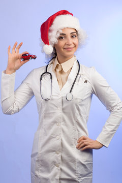 dottoressa in camice bianco , Stetoscopio intorno collo e con il cappello da babbo natale, mostra una machinetta giocattolo rossa,  isolata su sfondo celeste