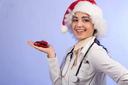 dottoressa in camice bianco , Stetoscopio intorno collo e con il cappello da babbo natale, mostra una machinetta giocattolo rossa,  isolata su sfondo celeste