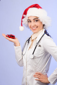 dottoressa in camice bianco , Stetoscopio intorno collo e con il cappello da babbo natale, mostra una machinetta giocattolo rossa,  isolata su sfondo celeste