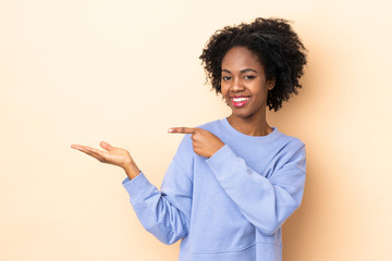 Young African American woman isolated on beige background holding copyspace imaginary on the palm...