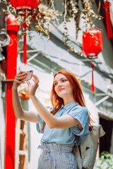Red hair tourist uses mobile phone for picture with smartphone of Yuyuan Garden street with red lanterns in Shanghai, china. Asia tourism travel. People taking photos during vacation lifestyle concept
