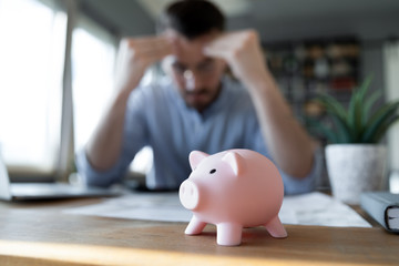 Close up unhappy man having financial problem, frustrated young male feeling anxiety about debt or bankruptcy, financial problem, lack of money, sitting at work desk with pink piggy bank