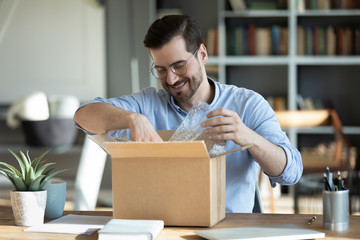 Smiling man wearing glasses unpacking awaited parcel, looking inside, sitting at work desk, satisfied happy customer opening cardboard box with online store order, good shipping delivery service - obrazy, fototapety, plakaty
