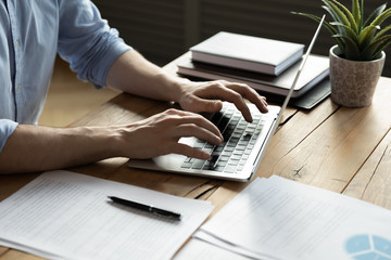 Close up businessman using laptop, typing on keyboard, sitting at wooden desk with documents,...