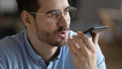 Close up head shot satisfied man wearing glasses holding smartphone near mouth, recording voice...