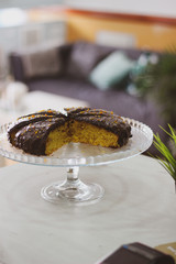 sliced orange cake with chocolate icing on glass stand with plant and sofa in the background