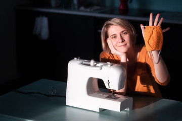 A tired young adult woman sits at a sewing machine and shows an orange mask sewn by her