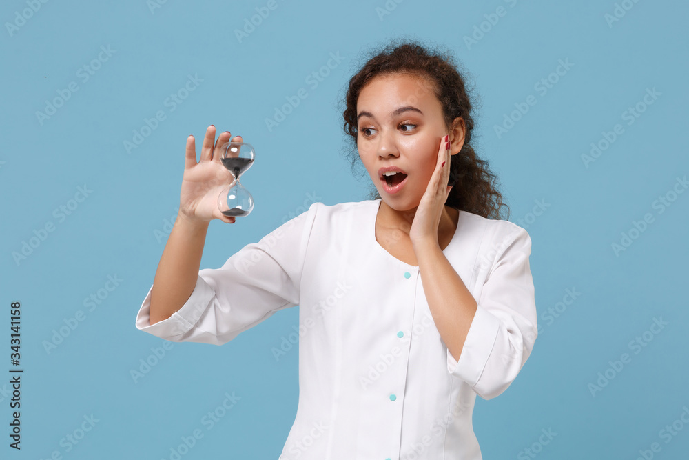 Wall mural Shocked african american doctor woman isolated on blue wall background. Female doctor in white medical gown hold hourglass black sand. Healthcare personnel medicine health concept. Mock up copy space.