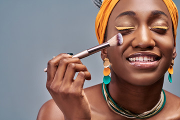 Young African woman in Studio doing body care and applying makeup, powder and rouge with a brush for young and beautiful skin