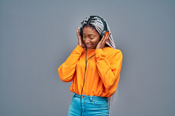 Adorable afro american woman in yellow shirt singing and having fun while listening to music using earphones isolated over gray background