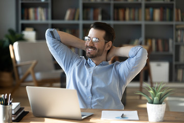 Smiling businessman freelancer wearing glasses leaning back in chair with hands behind head, happy satisfied young man dreaming about good future, new opportunities, visualizing success