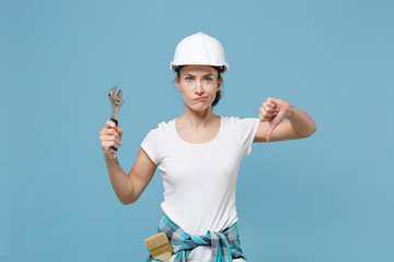Displeased young woman in protective helmet hardhat hold adjustable wrench isolated on blue...