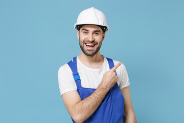 Funny young man in coveralls protective helmet hardhat isolated on blue wall background. Instruments accessories for renovation apartment room. Repair home concept. Pointing index finger aside up.