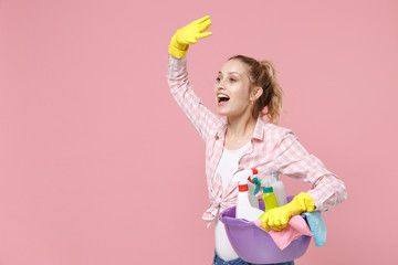 Cheerful young woman housewife in rubber gloves hold basin with detergent bottles washing cleansers while doing housework isolated on pink background studio. Housekeeping concept. Showing victory sign