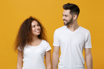 Smiling young couple two friends european guy african american girl in white t-shirts posing isolated on yellow background studio. People lifestyle concept. Mock up copy space. Looking at each other.