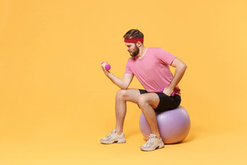Fototapeta na wymiar Tensed bearded fitness sporty guy 20s sportsman in headband t-shirt in home gym isolated on yellow background. Workout sport motivation lifestyle concept. Sit on fitball doing exercise with dumbbell.