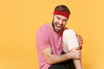 Ingured screaming young bearded fitness sporty guy sportsman in headband t-shirt in home gym isolated on yellow background. Workout sport motivation concept. Sitting touch knee with elastic bandage.