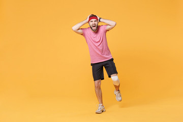 Fototapeta na wymiar Ingured screaming young bearded fitness guy sportsman in headband t-shirt in home gym isolated on yellow background. Workout sport motivation concept. Put hands on head with elastic bandage on knee.
