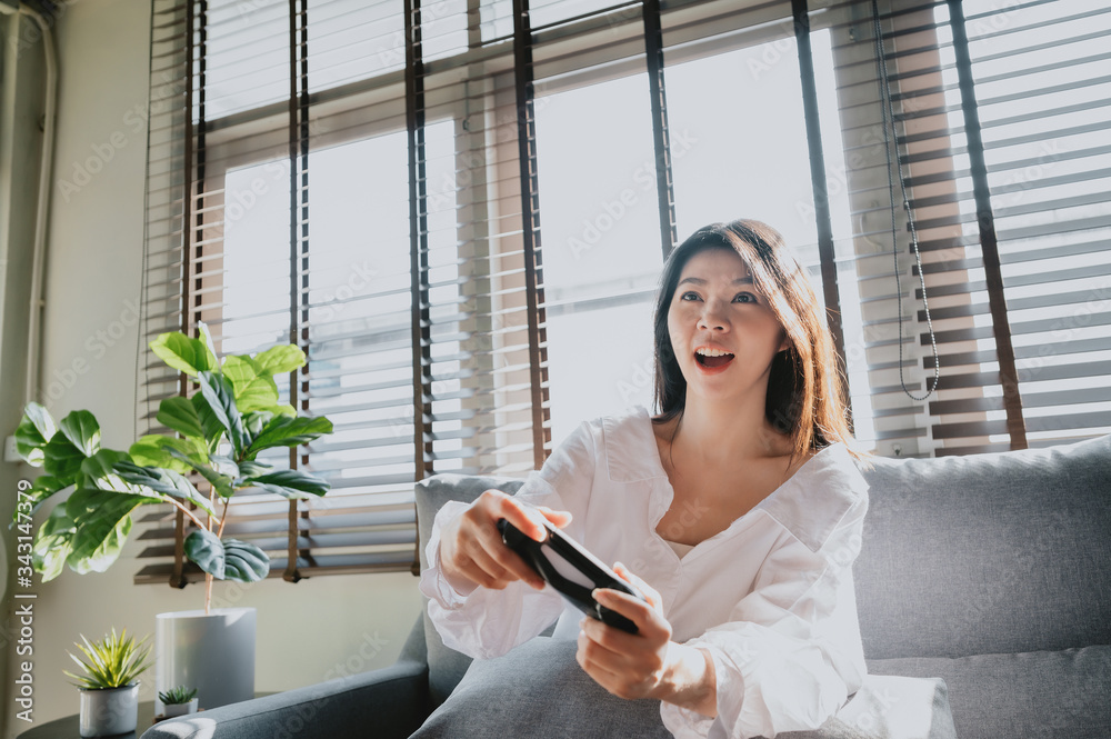 Wall mural Excited Asian woman looking at tv screen and holding controller while playing video game at home in living room