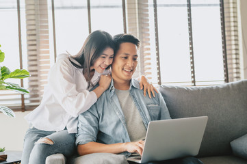 Happy Asian couple using laptop on sofa at home. Woman embracing man from behind
