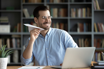 Happy smiling businessman wearing glasses playing with paper plane at workplace, sitting at desk,...