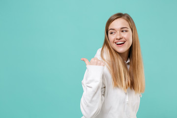 Side view of cheerful young woman girl in casual white hoodie posing isolated on blue turquoise background studio portrait. People emotions lifestyle concept. Mock up copy space. Pointing thumb aside.