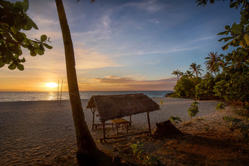 a white beach topical scene with sun on horizon and sunset reflection in sea water with a beach cottage, hut, at the Beach