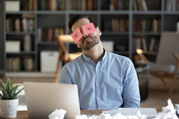 Exhausted tired businessman with painted eyes on stickers, adhesive notes on face sleeping at workplace, sitting at desk with laptop, unproductive lazy young male dozing, working on difficult project - obrazy, fototapety, plakaty