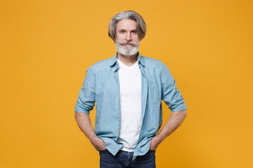 Elderly gray-haired mustache bearded man in casual blue shirt posing isolated on yellow background studio portrait. People emotions lifestyle concept. Mock up copy space. Holding hands in pockets.