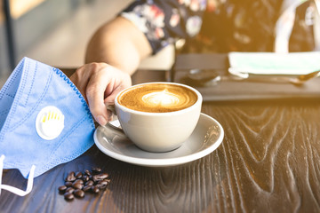 Woman with medical face mask drinking coffee in her house alone during corona virus or COVID-19 quarantine, Physical distancing for protect Covid-19 concept. Selective focus.