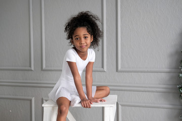A portrait of an African ballerina in a white gymnastic costume sits on a wooden chair.