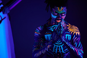 portrait of calm african man with fluorescent prints on skin praying for the best, stand in the pose of prayer. luminescence, fluorescence, body art concept