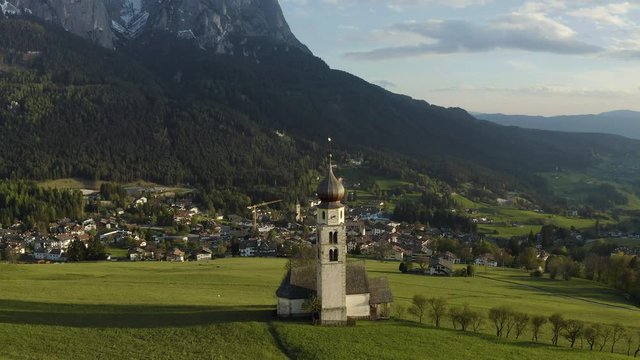 Aerial view of picturesque valley with chapel in Bolzano at sunset, Trentino, amazing green meadows of the mountains of Italy, roof tops of houses, Dolomites on background, drone flies around chapel