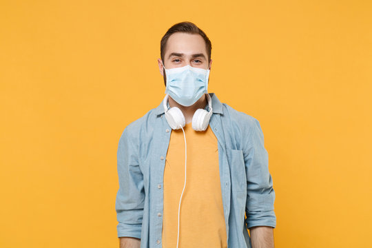 Cheerful young man in sterile face mask with headphones posing isolated on yellow background studio portrait. Epidemic pandemic rapidly spreading coronavirus 2019-ncov sars covid-19 flu virus concept.