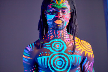 portrait of young black man with colorful ethnic fluorescent prints on his muscular body, shirtless guy posing at camera, colorful makeup and body art - obrazy, fototapety, plakaty