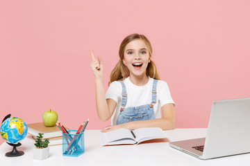 Excited little kid schoolgirl 12-13 years old sit study at white desk with pc laptop isolated on pink background. School distance education at home during quarantine concept. Pointing index finger up.