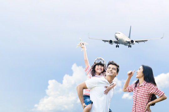 Father Carry Boy Wearing Captain Hat On Back, Playing Airplane Toy In Sky Together With Mother. Young Child Want To Be Pilot In Future When He Growing Up. Family Supports Son Dream Job Or Occupation.