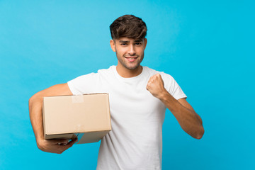 Young handsome man over isolated blue background holding a box to move it to another site