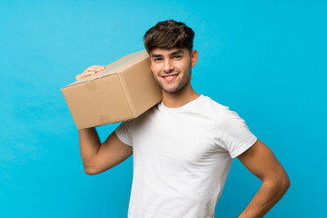 Young handsome man over isolated blue background holding a box to move it to another site
