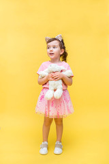 Cute girl with tails and cat ears on a yellow background. A little girl holds a rabbit toy. White rabbit toy. A girl in a pink t-shirt and skirt. Sideways view. Copy space.