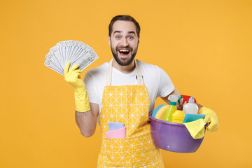 Surprised young man househusband in apron rubber gloves hold basin with detergent bottles washing...