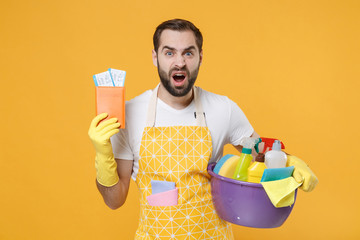 Irritated young man househusband in apron rubber gloves hold basin with detergent bottles washing...