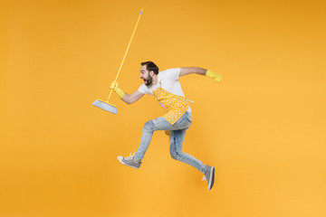 Side view of crazy young man househusband in apron rubber gloves hold in hands broom while doing housework isolated on yellow wall background studio portrait. Housekeeping concept. Jumping, screaming.